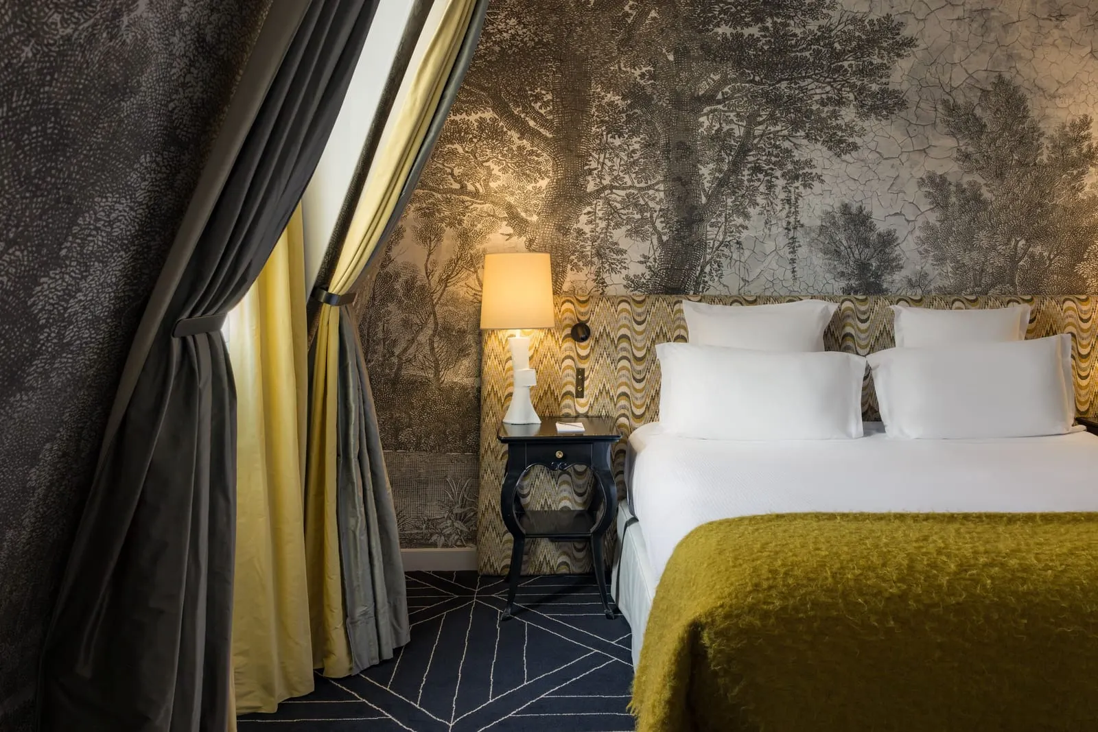 Connecting rooms (two Superior Rooms) at Le Pavillon de la Reine: one of the bedrooms, with a double bed and an olive throw against a tree mural, and curtains drawn back from a window in a sloping wall