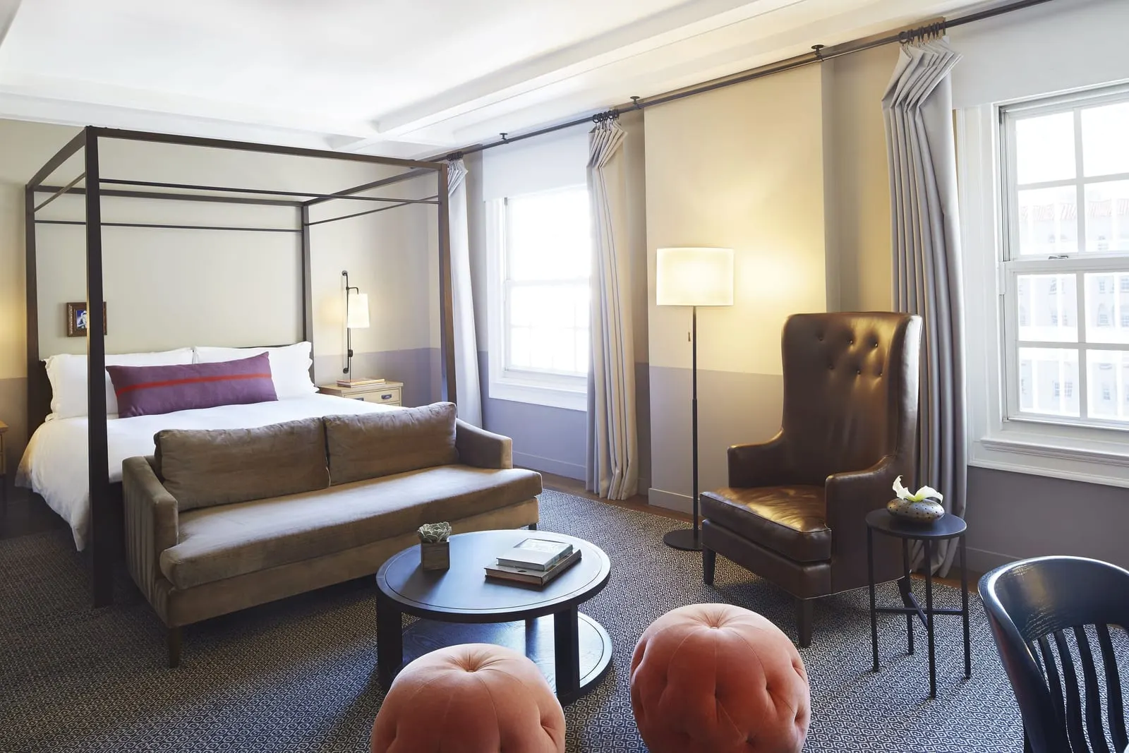 Premiere Room at The Hollywood Roosevelt: a four-poster bed with a velvet sofa at its foot, a round coffee table, two pink poufs and a leather armchair by tall sash windows