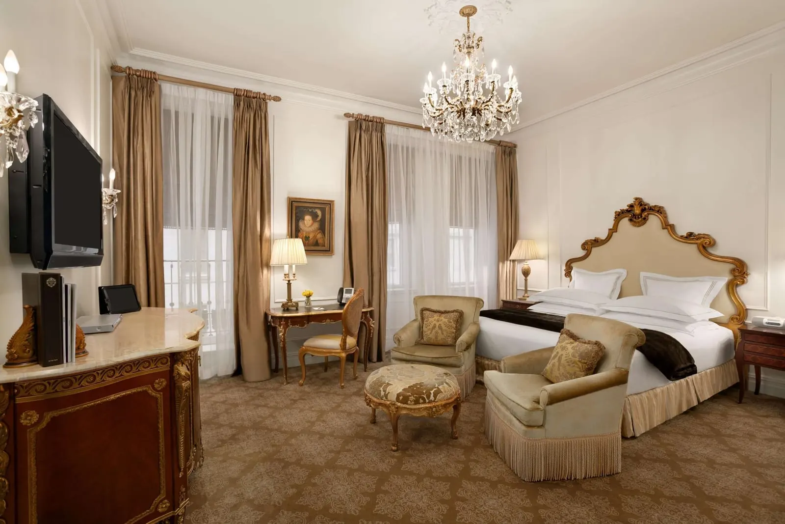 Terrace Room King at The Plaza: a king bed with a gilded headboard under a crystal chandelier, a wall mounted television, a writing desk and two tall windows with sheer curtains