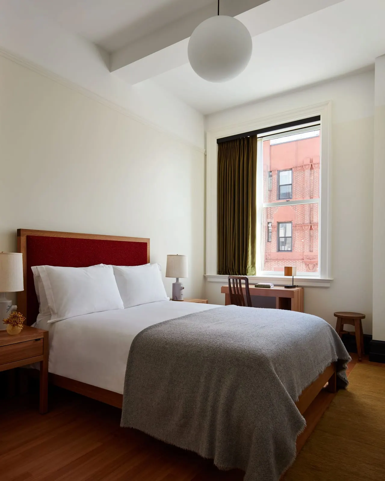 Studio Queen at Nine Orchard: a bed with a red upholstered headboard and a grey wool throw beside a window with an olive curtain, a small wooden desk below the window