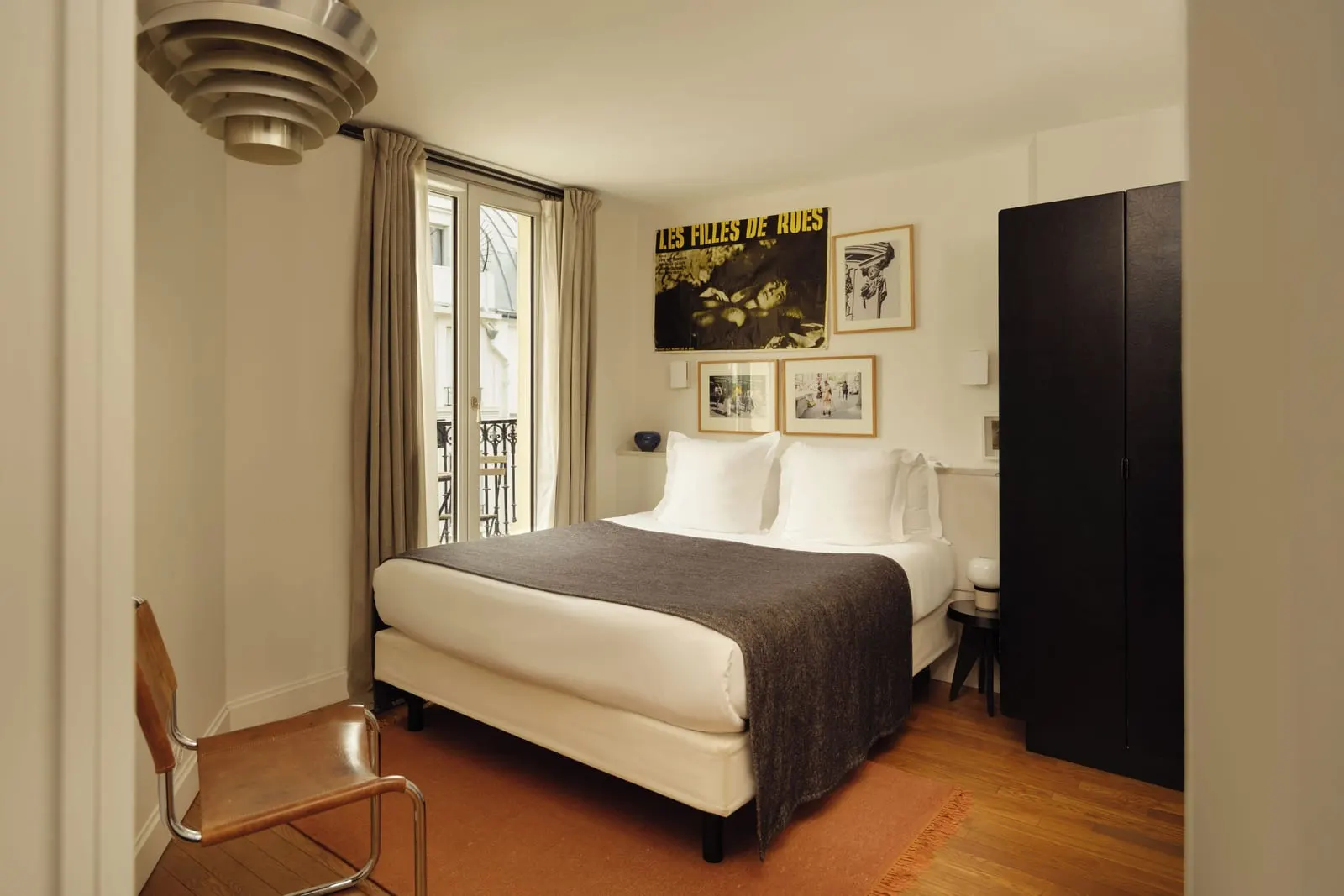 Pigalle 17 at Le Pigalle: a double bed under a vintage film poster and framed photographs, a tall black wardrobe, a tubular leather chair and a French window with a wrought-iron railing