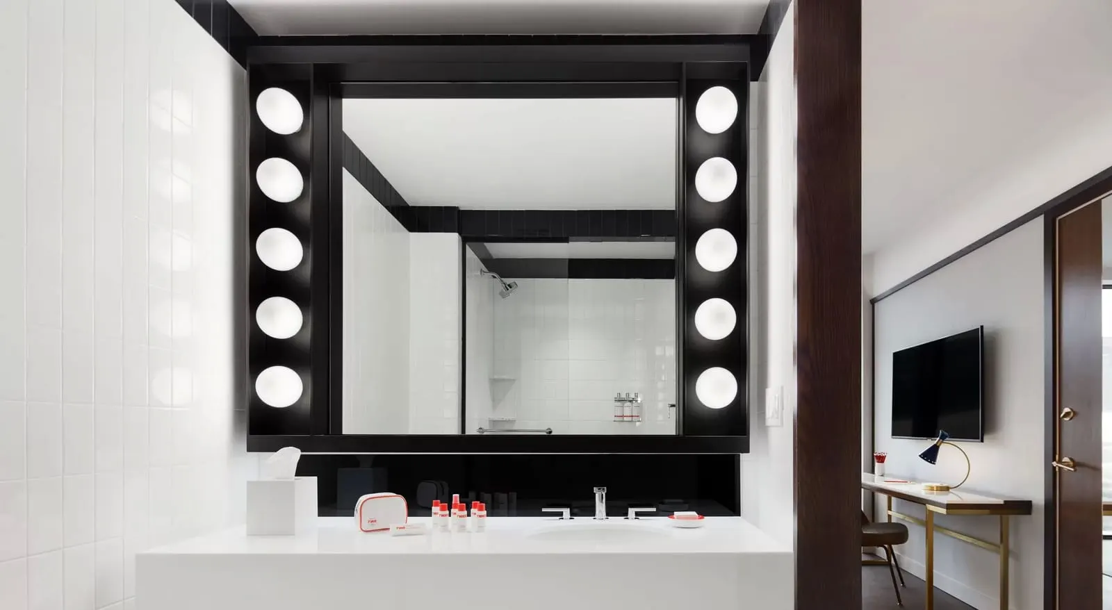 Deluxe King With Historic TWA View at TWA Hotel: a bathroom vanity under a mirror framed by a column of round bulbs on each side, with the shower reflected in the glass and the desk and television visible through the doorway