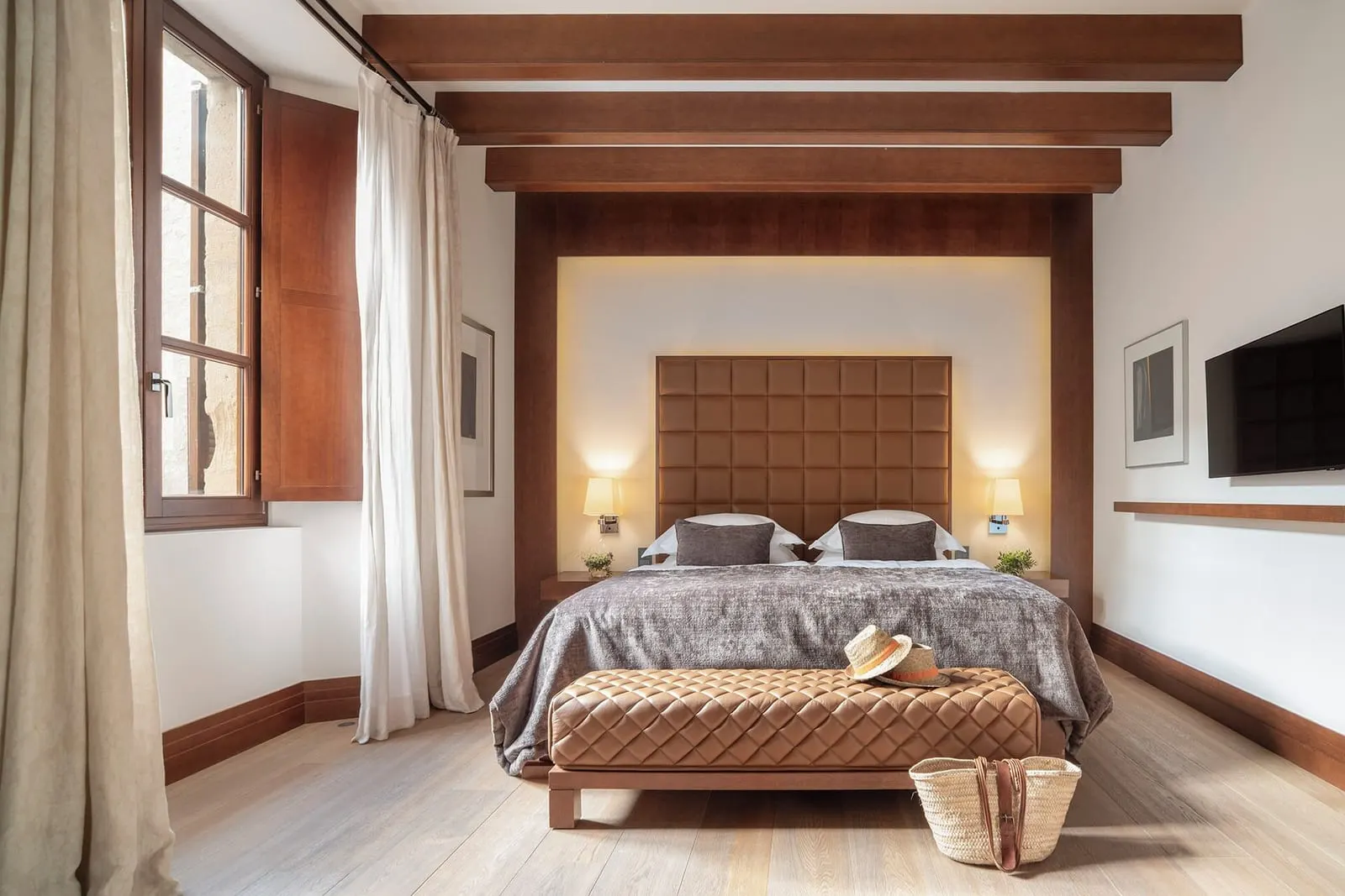 Estate Double im Castell Son Claret: ein Bett mit gestepptem Kopfteil und einer Lederbank unter dunklen Holzbalken, ein Wandfernseher und ein Fenster mit Fensterläden und langen Vorhängen
