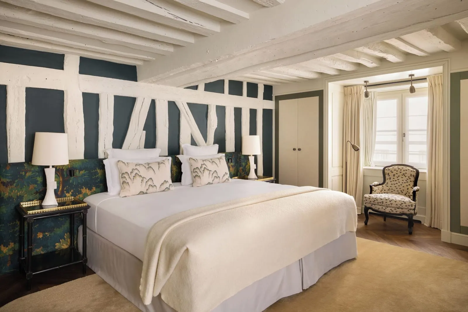 Family Suite at Le Pavillon de la Reine: a bedroom with a double bed against a wall painted with timber framing, whitewashed beams, a wardrobe and an armchair by the window