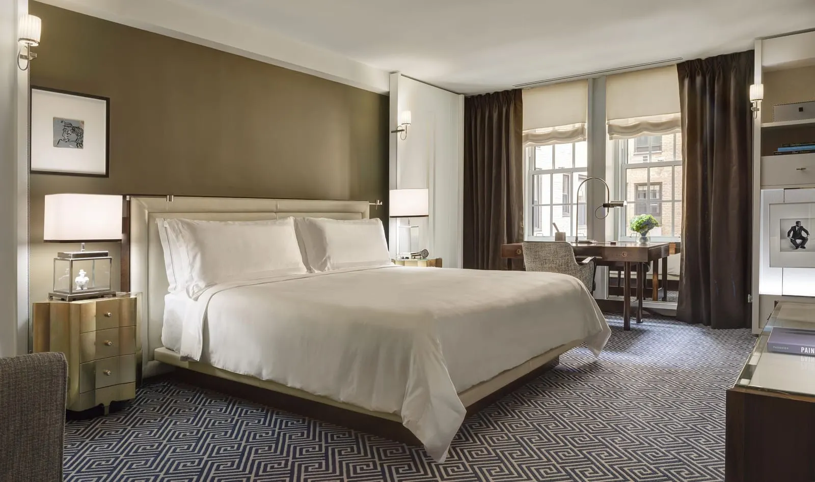 Deluxe King Room at The Carlyle: a king bed beside a writing desk set under tall windows, with taupe walls and heavy brown curtains