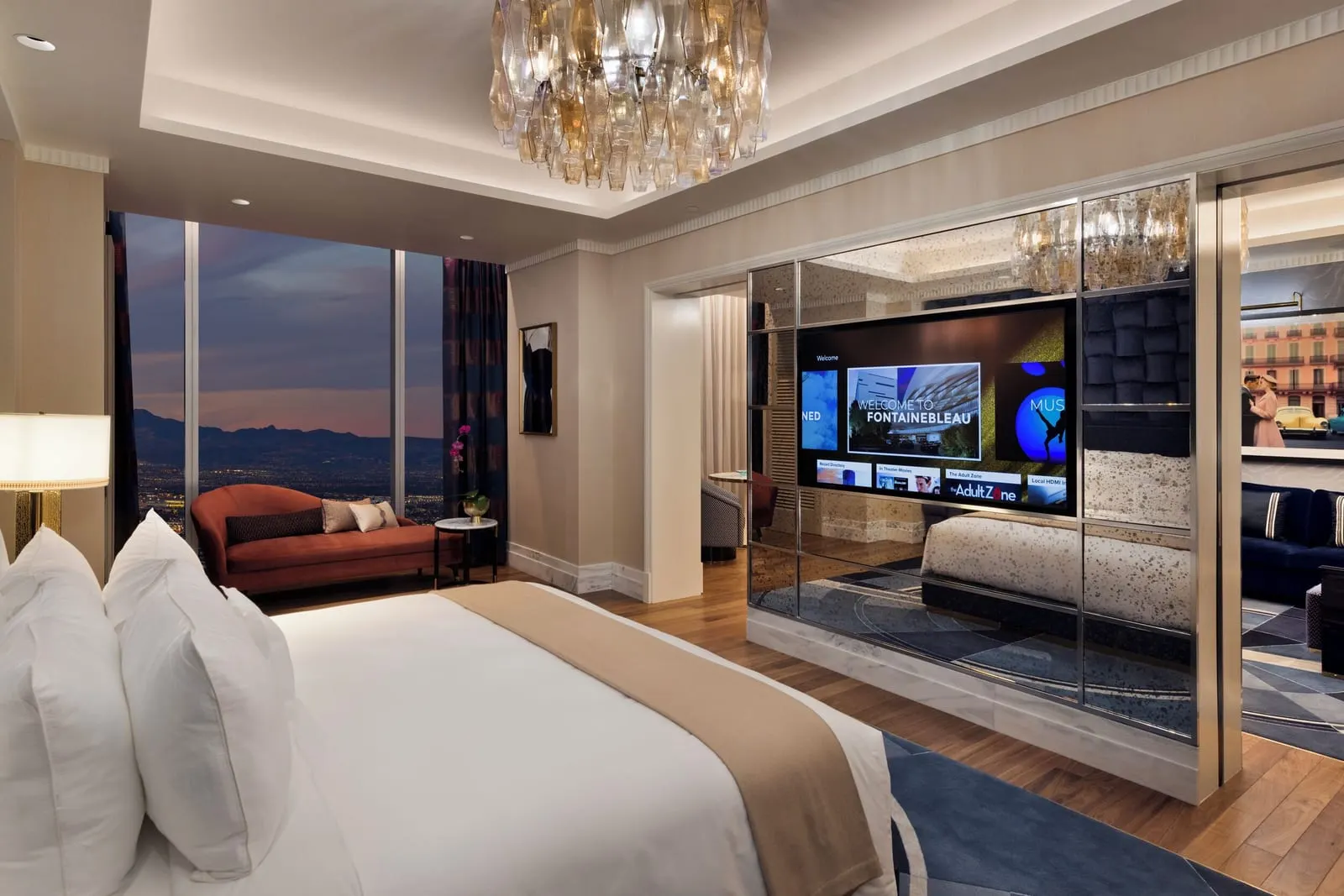 Fleur de Lis Monogram Suite at Fontainebleau Las Vegas: a king bed under a glass chandelier, a mirrored partition holding a television, and a rust-coloured chaise longue by floor-to-ceiling windows