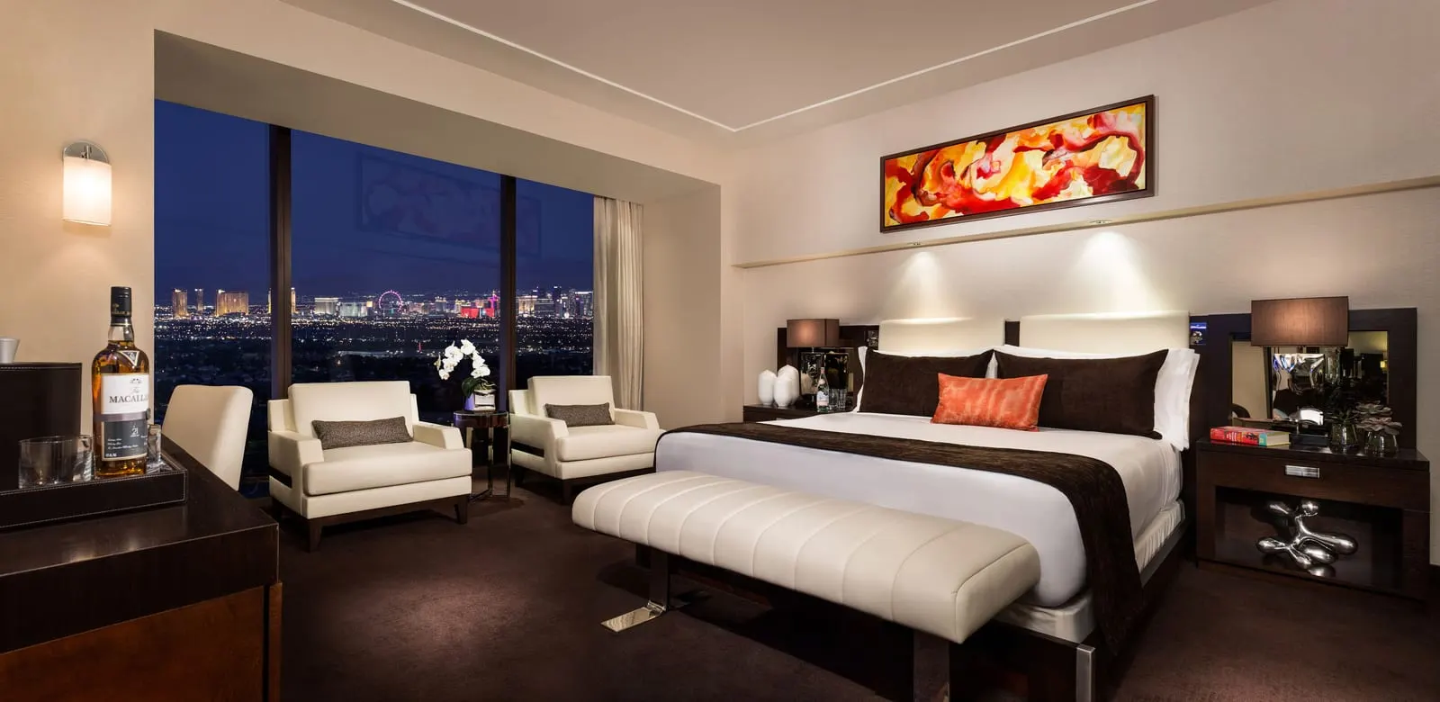 Two Bedroom Luxury Suite at Red Rock Casino Resort & Spa: a bedroom with a bed below an orange and red abstract painting, a cream bench at its foot and two cream armchairs by the window