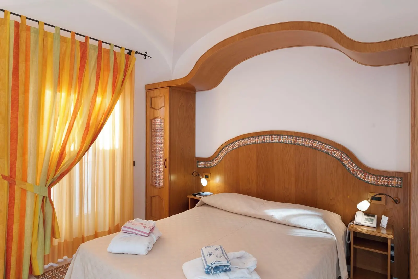 Superior Room with balcony and partial sea view all'Hotel Tramonto d'Oro: un letto matrimoniale con testiera in legno bordata di piastrelle in ceramica sotto un baldacchino ricurvo in legno, accanto a tende a righe arancioni.