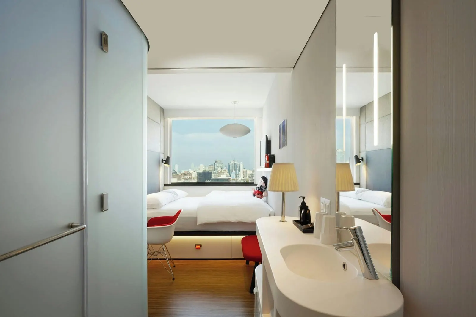 Guest room, 1 King, Premium view at citizenM New York Bowery: a king bed under a wide window that looks out over rooftops and towers, seen past the basin, a table lamp and the frosted shower pod