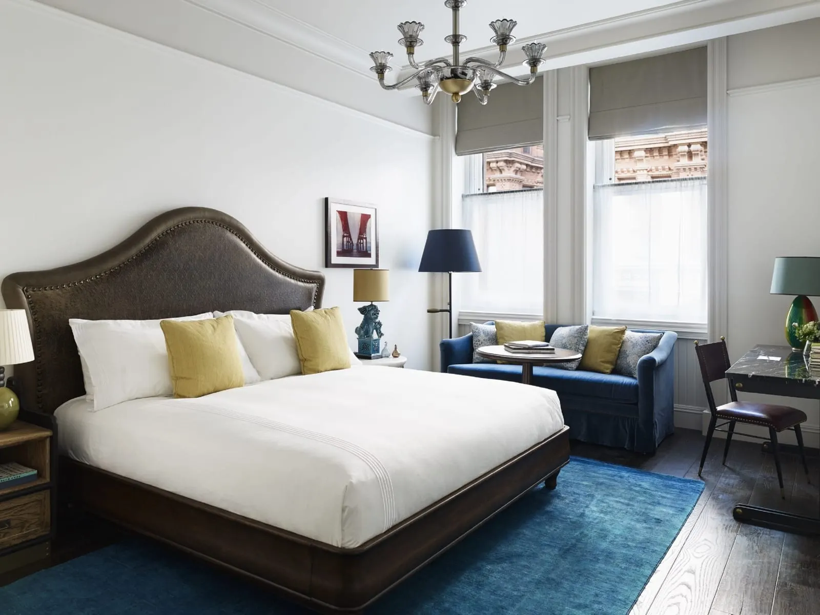 Premium King at The Beekman: a king bed on a blue rug, a blue sofa and round table in front of two tall windows, a dark marble desk to the right and a chandelier above