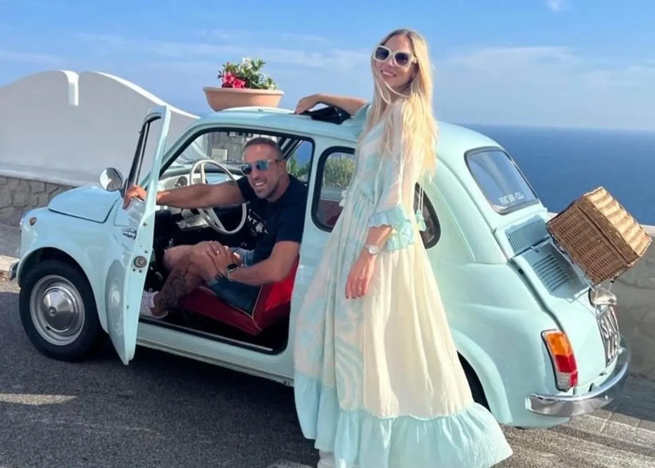 A pale blue vintage Fiat 500 with its door open and a wicker basket on the back, parked on a terrace above the sea, a man at the wheel and a woman in a long dress beside it