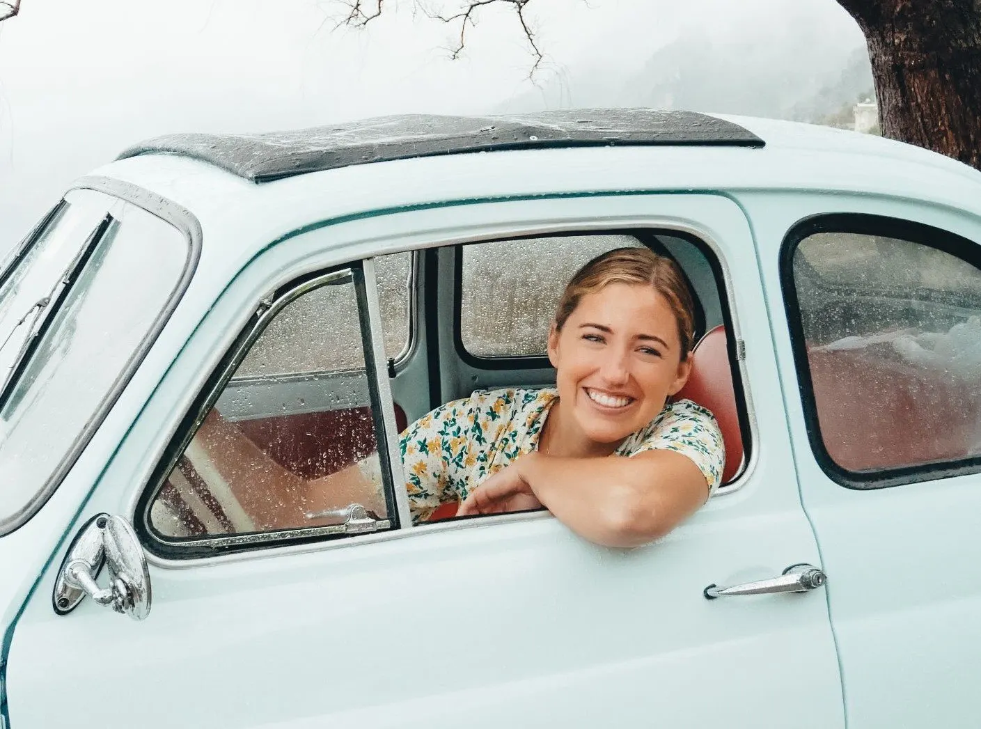 A smiling woman leaning out of the window of a pale blue vintage Fiat 500 with red seats, on a misty, rainy day on the coast