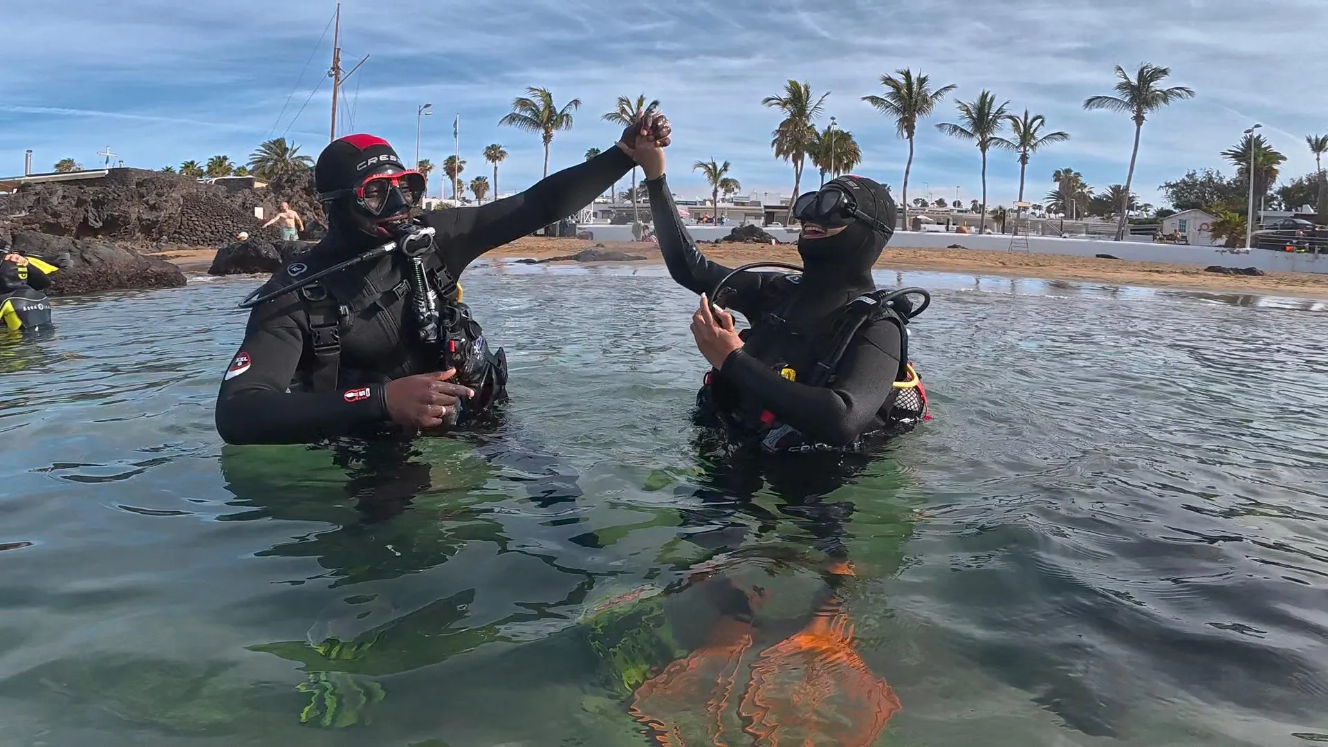 椰子の木と遊歩道を背に、腰の深さの穏やかな海で、スキューバ装備の2人のダイバーが手をつないで上げている様子