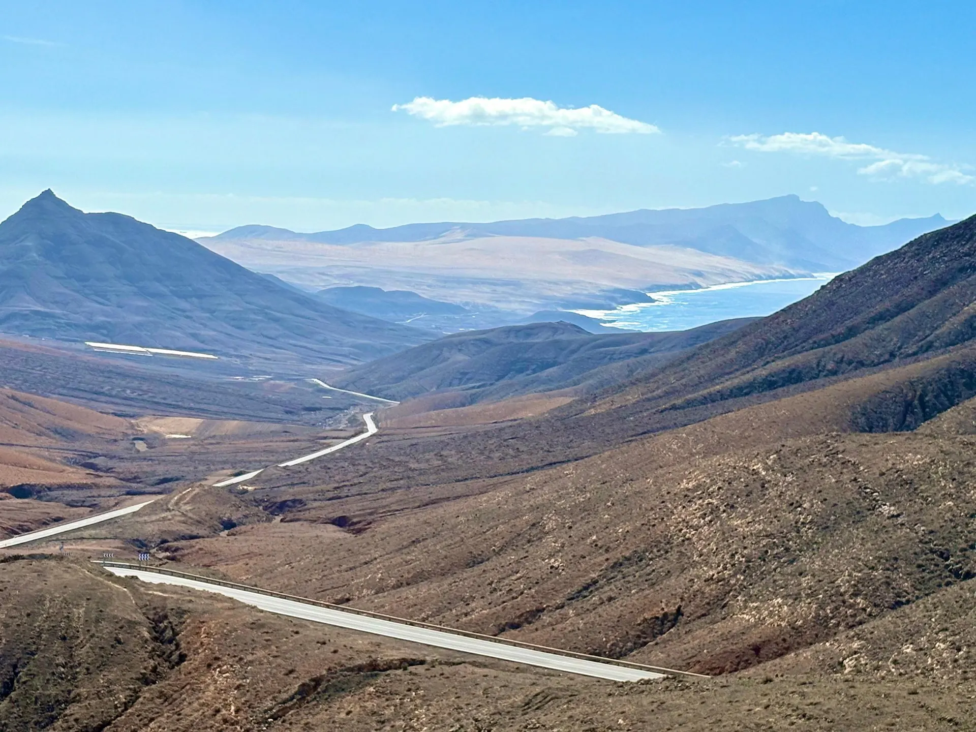 A road winding through bare brown hills towards the sea on Fuerteventura, under a pale blue sky