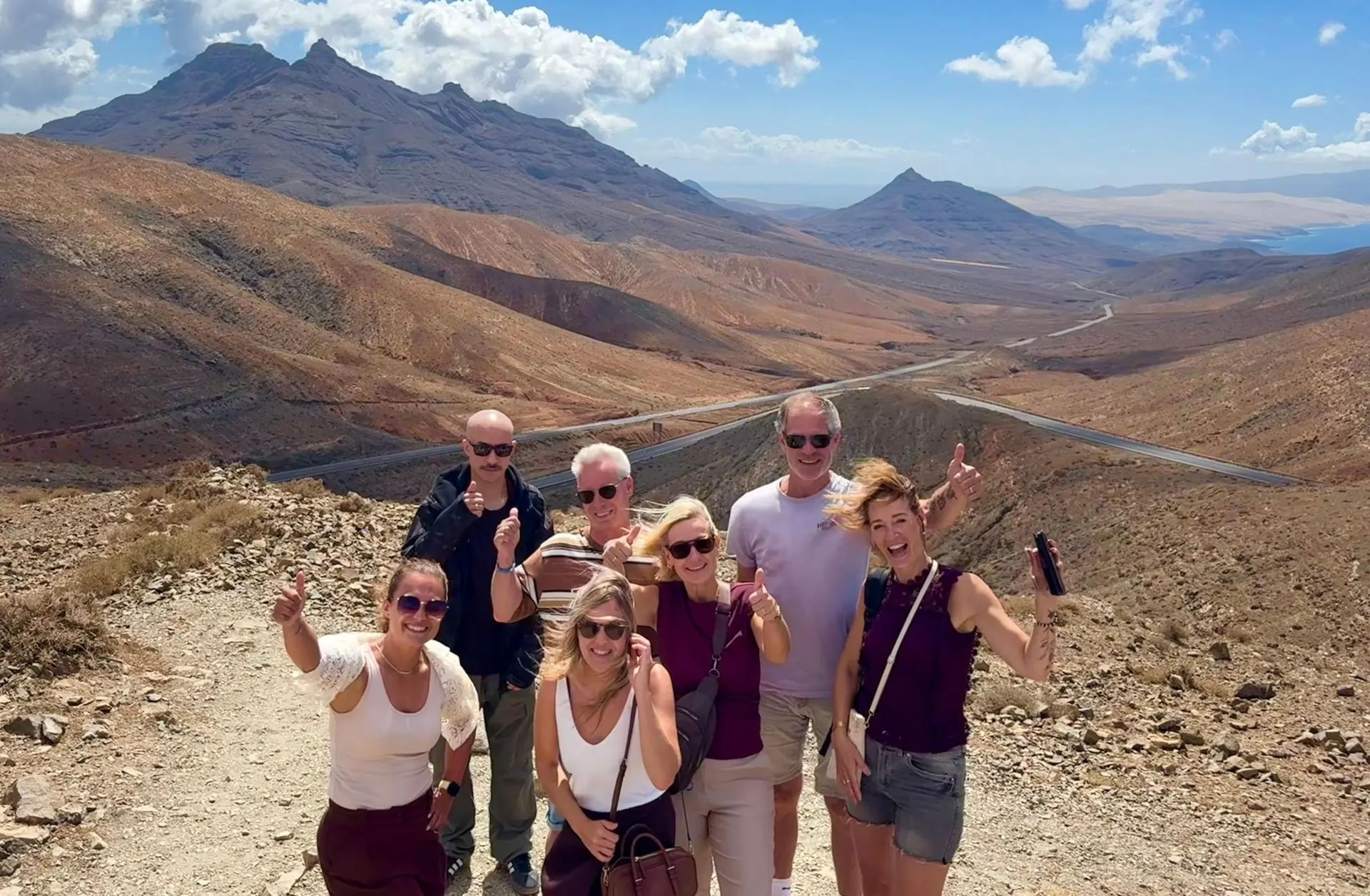 A small tour group posing on a mountain track above a valley road, with volcanic peaks and the coast behind