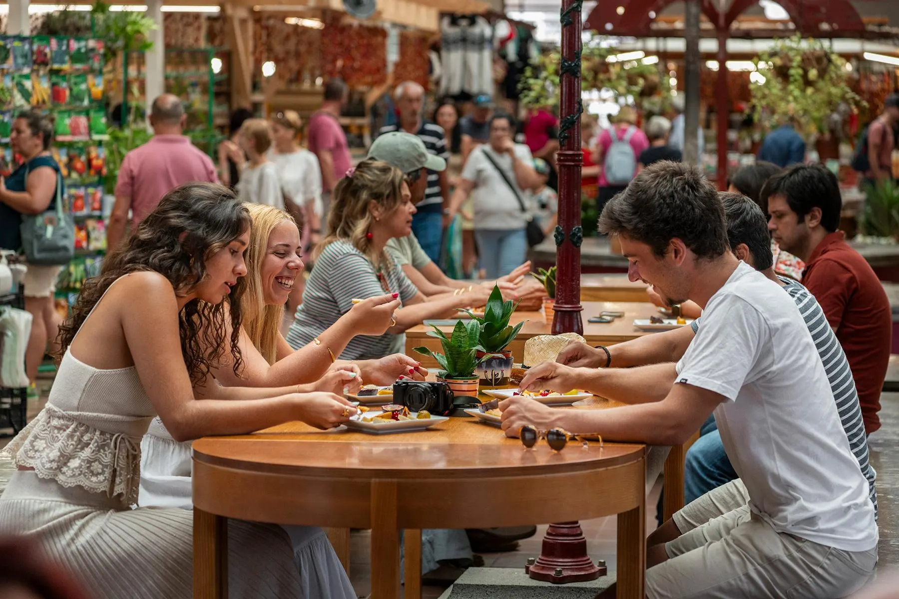 Visitors at wooden tables in the Mercado dos Lavradores in Funchal, eating plates of cut fruit, with market stalls and shoppers behind