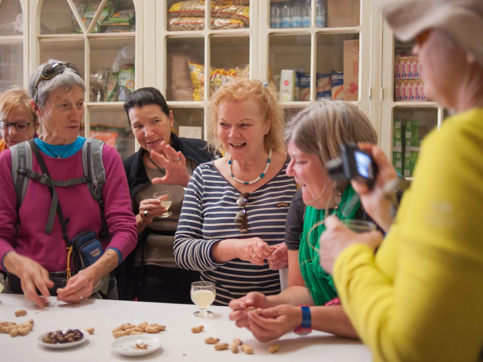 A tour group laughing round a counter in an old grocery shop, with small glasses of poncha and peanuts in their shells
