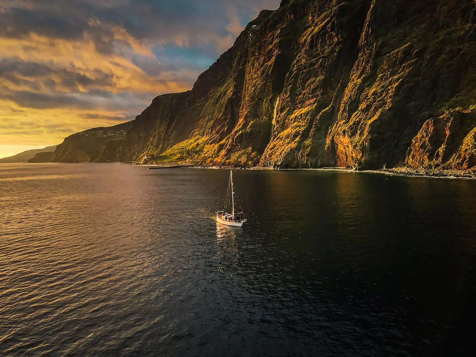 A small sailing yacht on calm water below tall green sea cliffs, in the low light of the evening