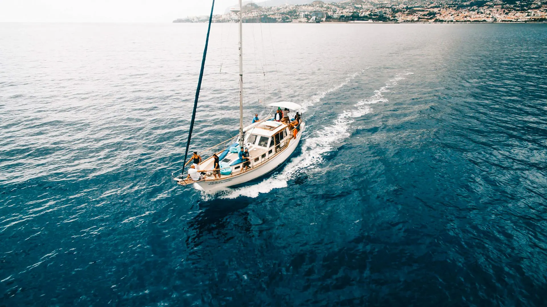 A wooden-trimmed sailing yacht with guests on deck, heading out from Funchal, with the city on the hillside behind
