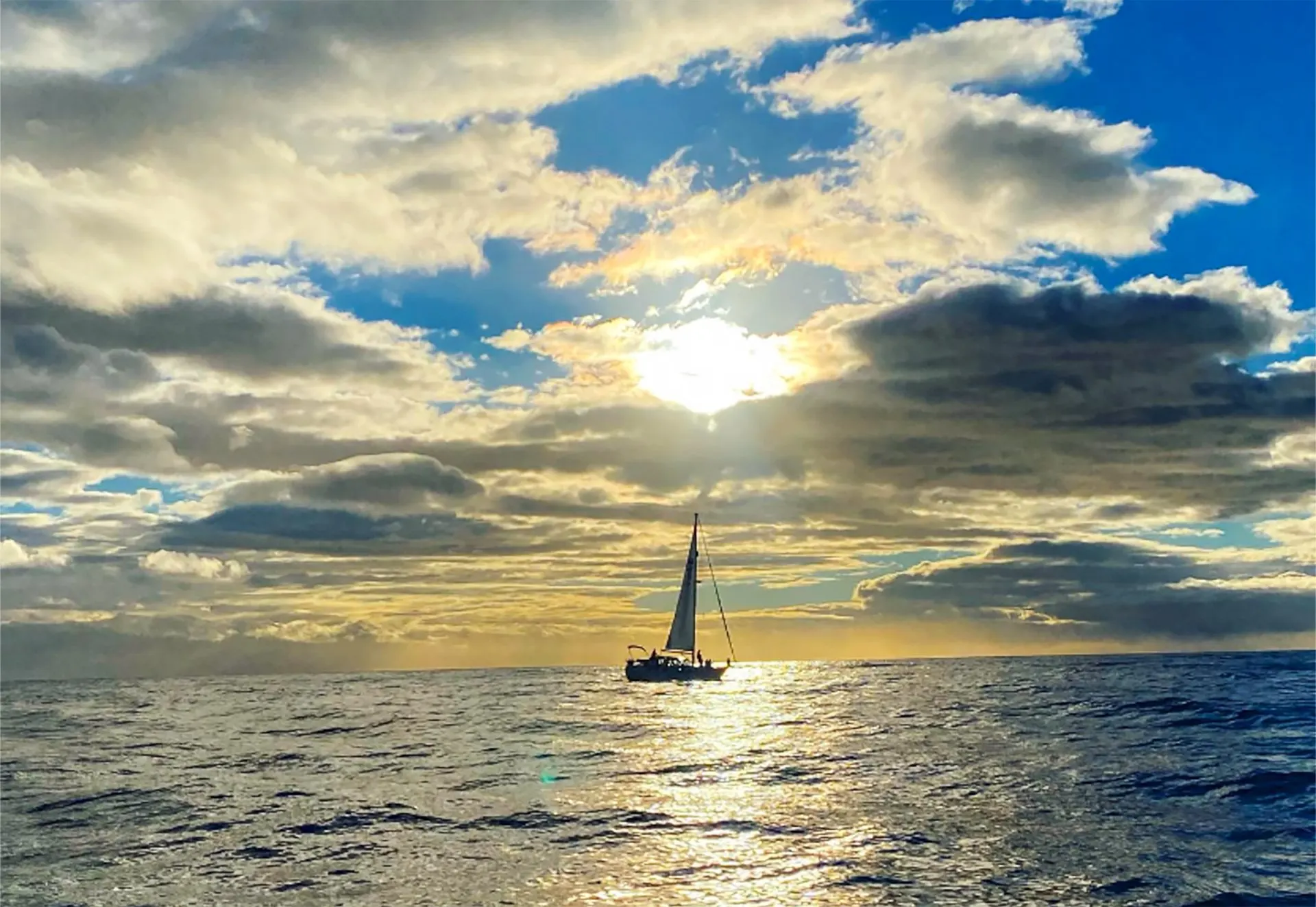 A yacht under sail in silhouette on open sea, under broken cloud with the sun behind it