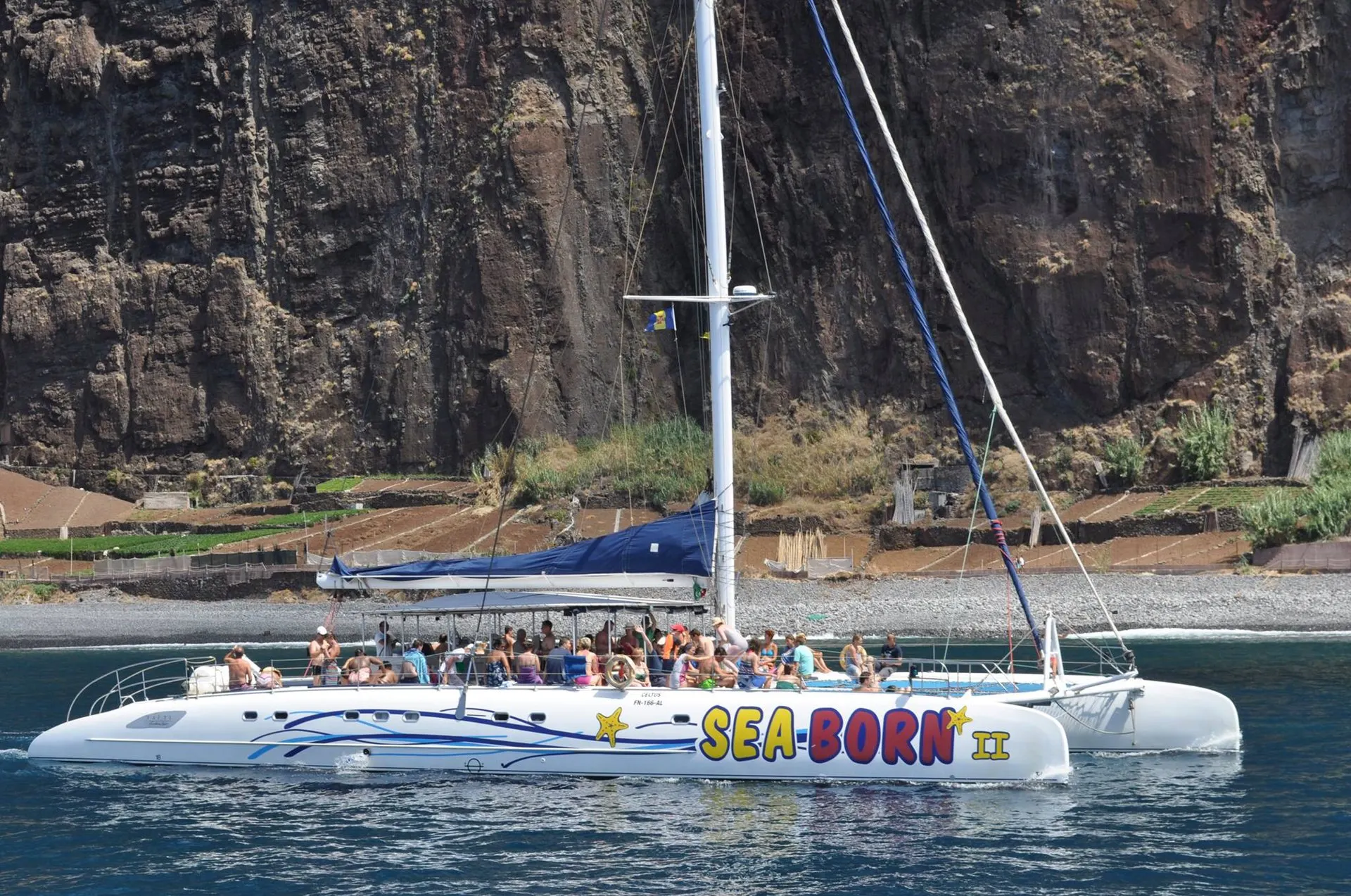 The white Sea Born II catamaran under sail beneath the sea cliffs west of Funchal, with passengers on deck