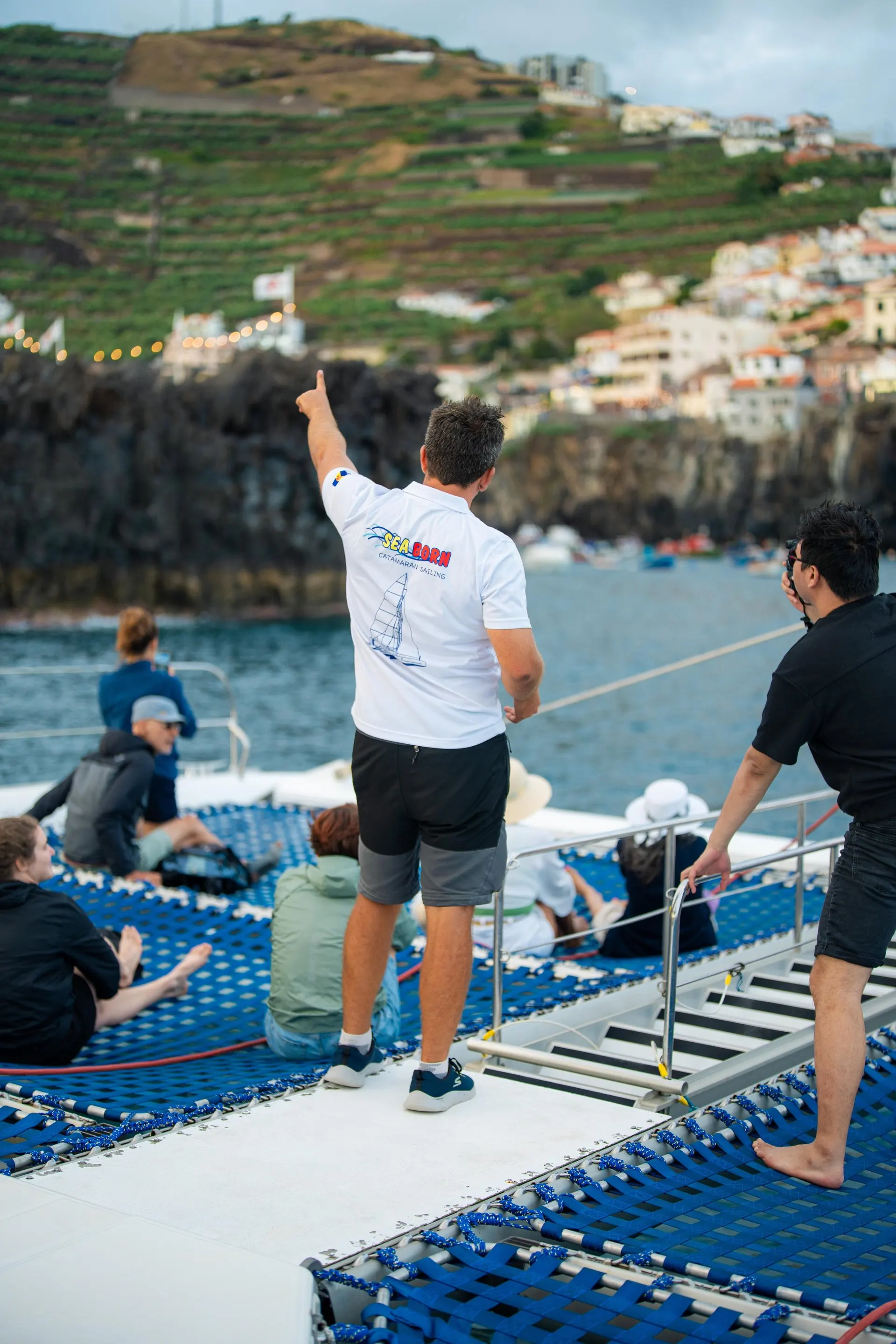 A crew member pointing out to sea from the catamaran's netting, with Câmara de Lobos behind