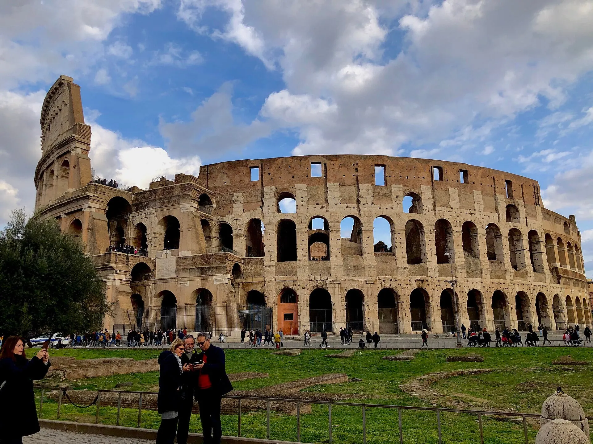 The Colosseum from the grass of the Colle Oppio side, its outer ring of arches against a blue sky