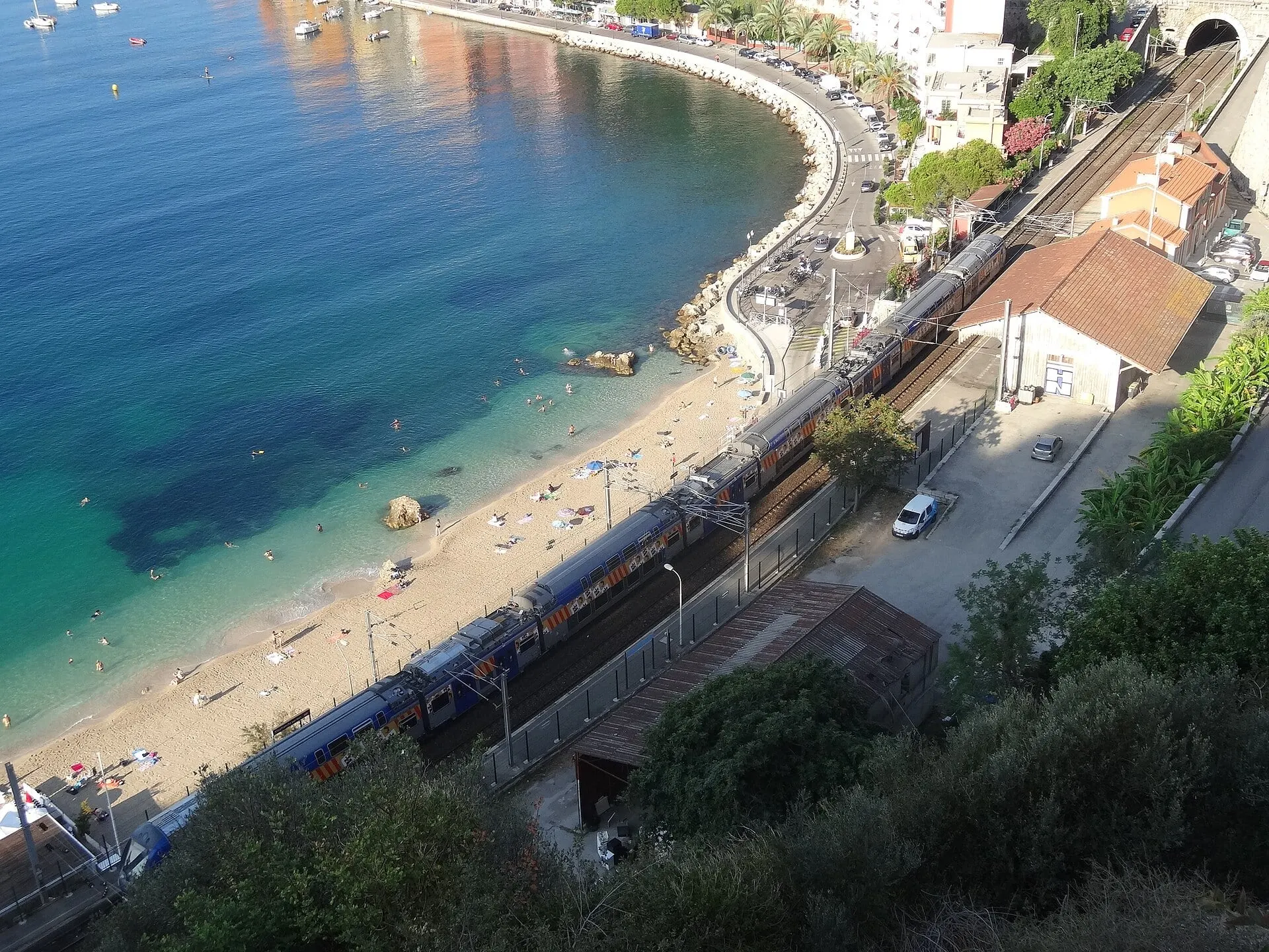 A double-deck regional train at Villefranche-sur-Mer station above the turquoise bay and its beach.