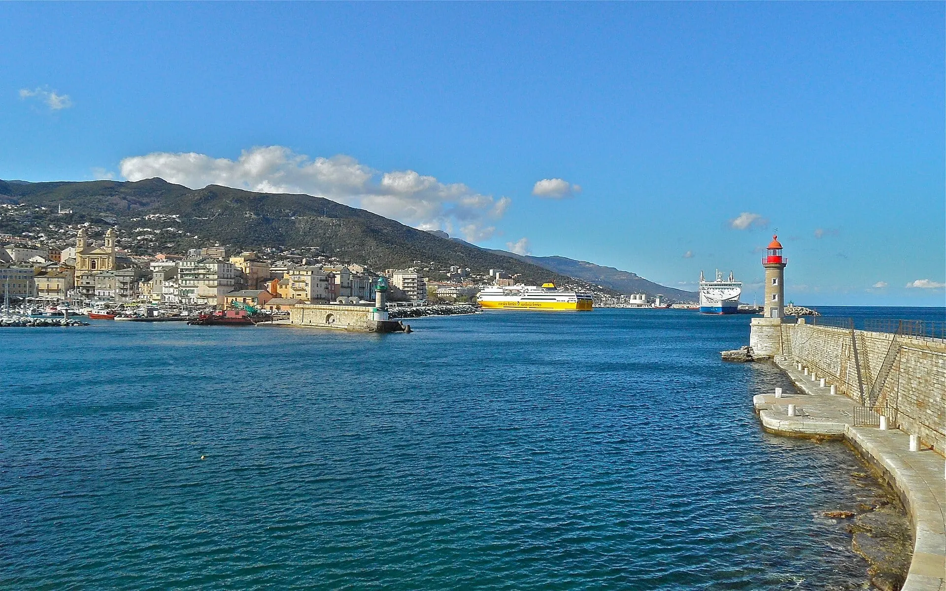 A yellow Corsica Ferries ship in the port of Bastia under a blue sky, seen from the old harbour jetty and its red lighthouse