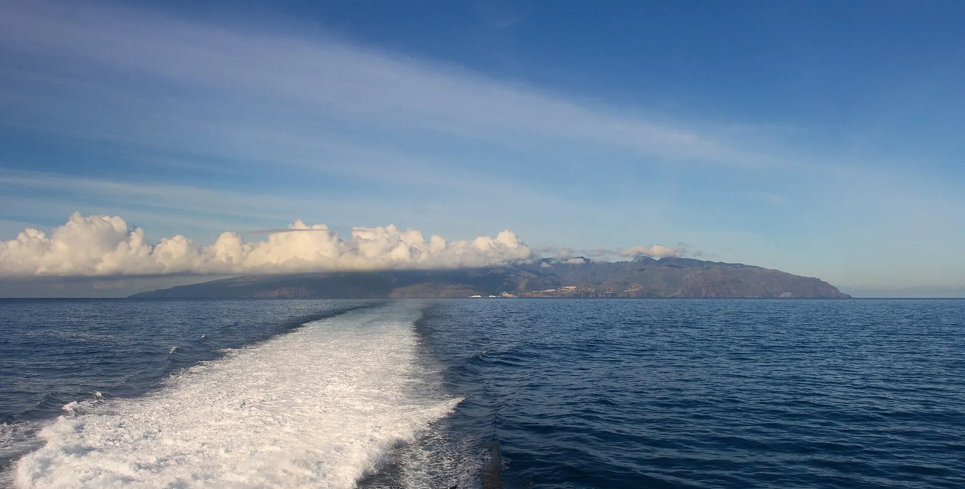 A ferry's white wake across the deep blue sea on the crossing between La Gomera and Tenerife, with the east coast of La Gomera under a bank of cloud.