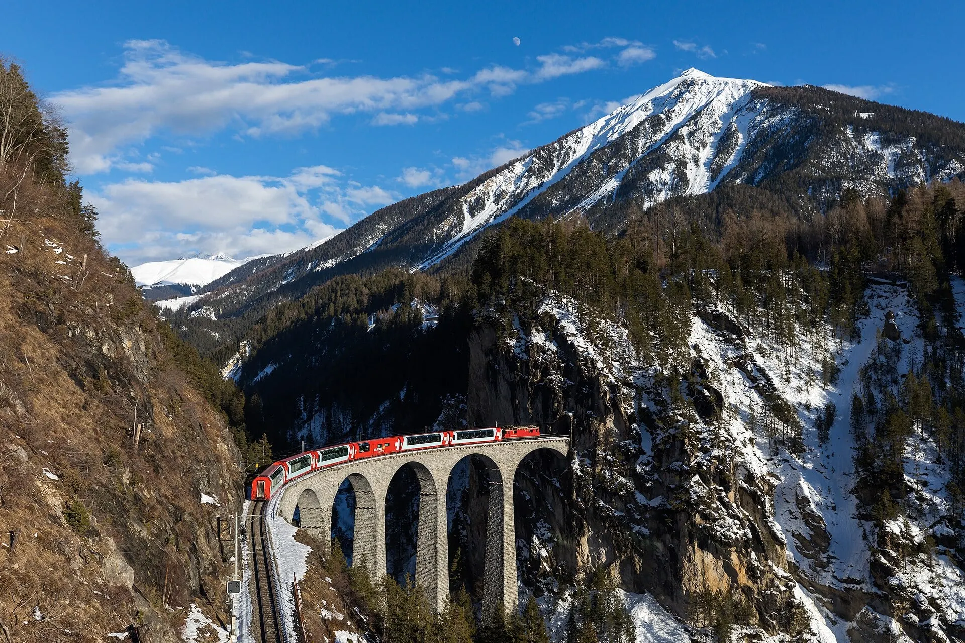 The Glacier Express crossing the Landwasser Viaduct under a snow capped peak and blue sky