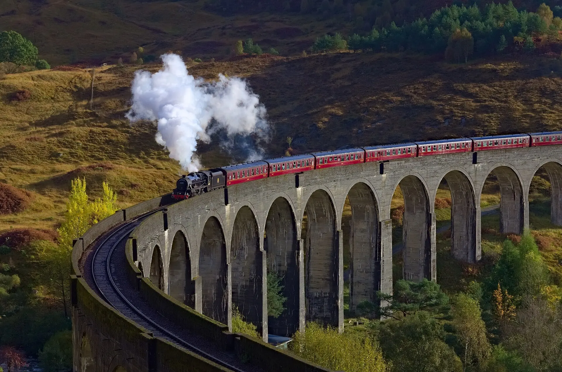 The Jacobite steam train crossing the curving Glenfinnan Viaduct in golden autumn light.