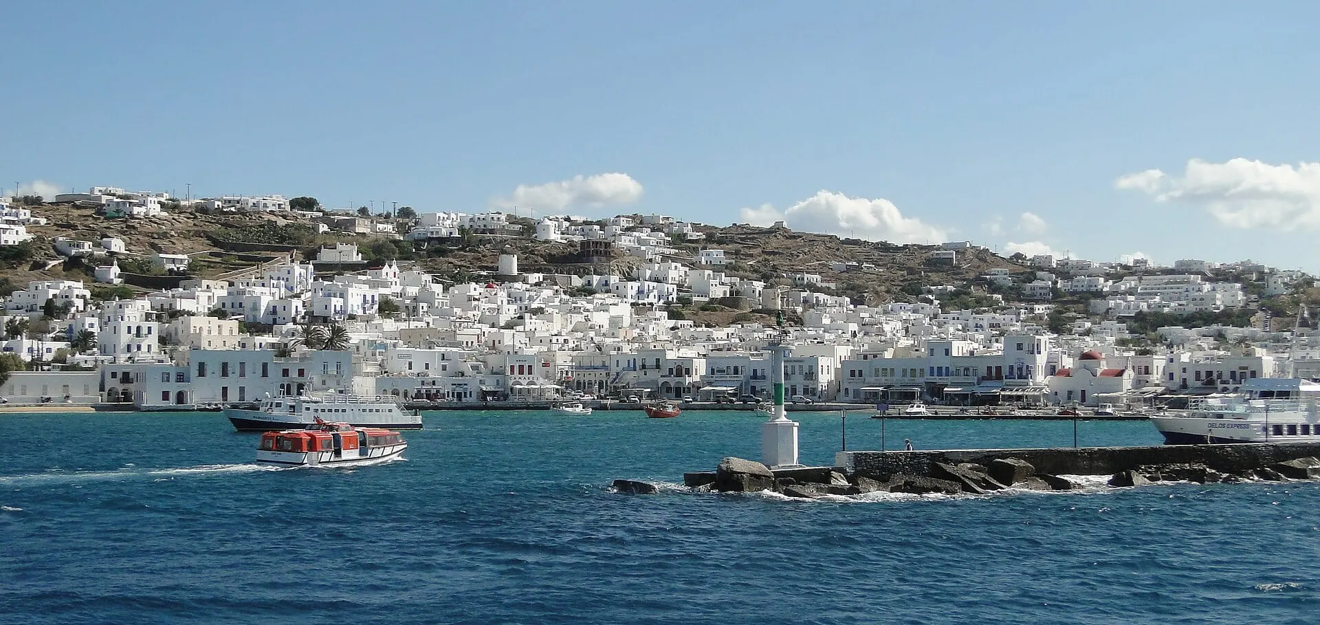The white town of Mykonos spread along the bay above the old harbour, with a small boat crossing the blue water