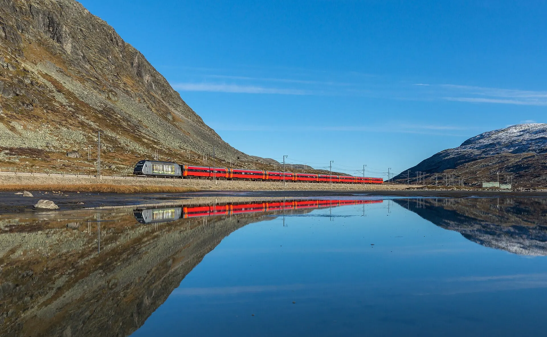 A Bergen Line train with red coaches reflected in still water under a blue sky on Hardangervidda between Haugastøl and Finse.