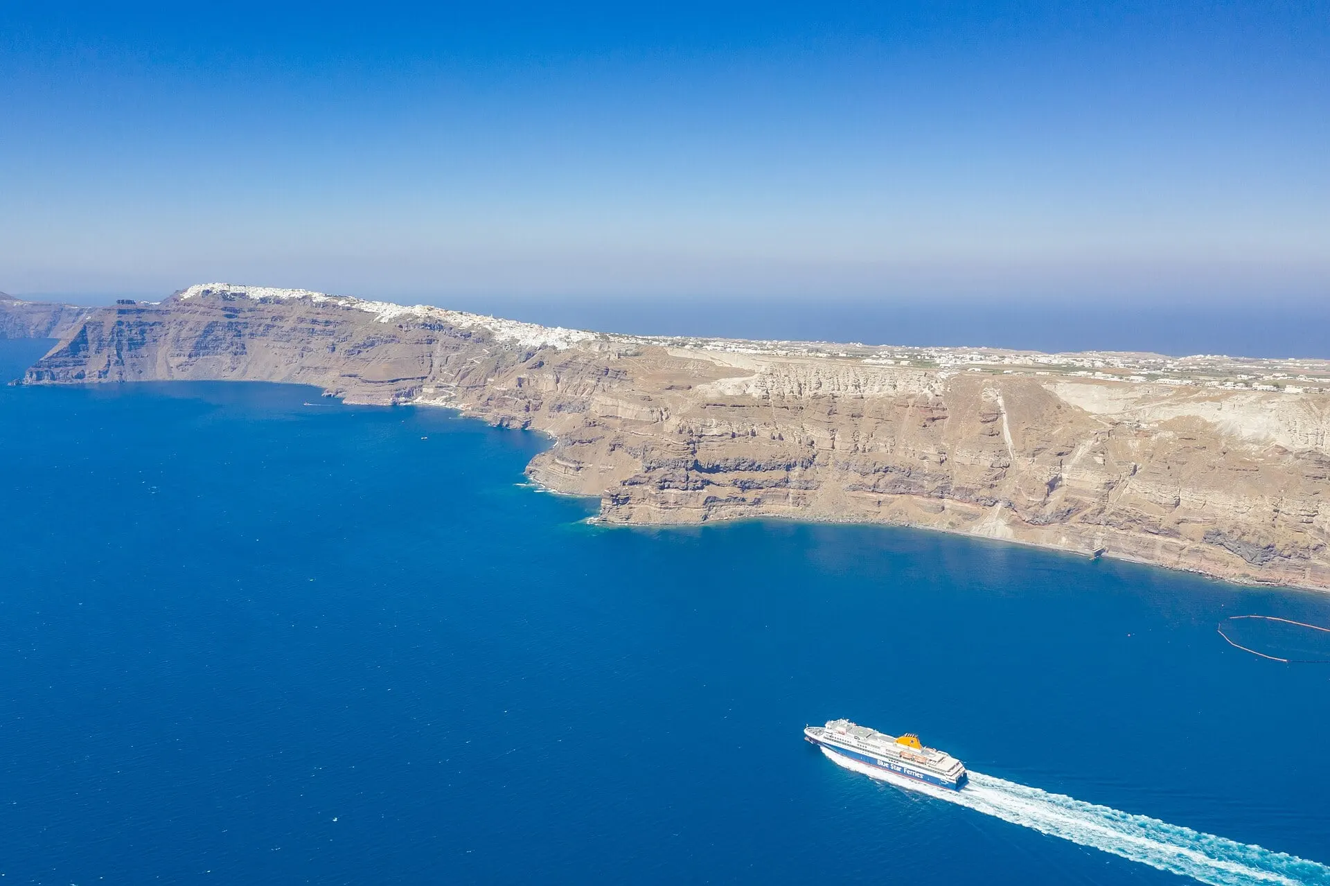 A Blue Star ferry crossing the deep blue Santorini caldera below the cliffs, with white villages along the rim.
