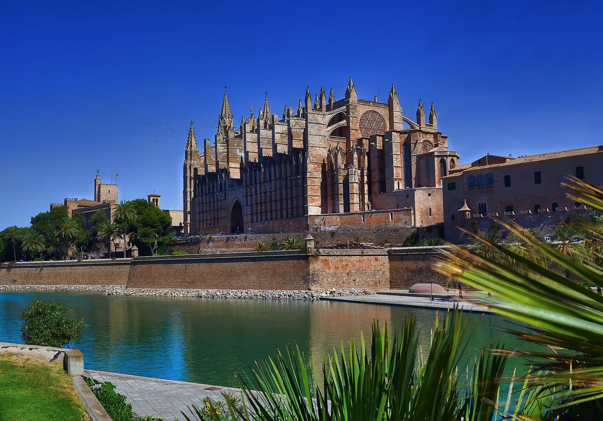 La Seu, the cathedral of Palma de Mallorca, above the old sea wall and the water of the Parc de la Mar under a deep blue sky.