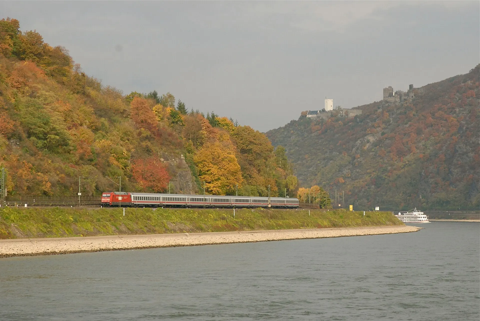 An Intercity train runs beside the Rhine under autumn woods, with the castles of the Hostile Brothers on the hillside beyond.