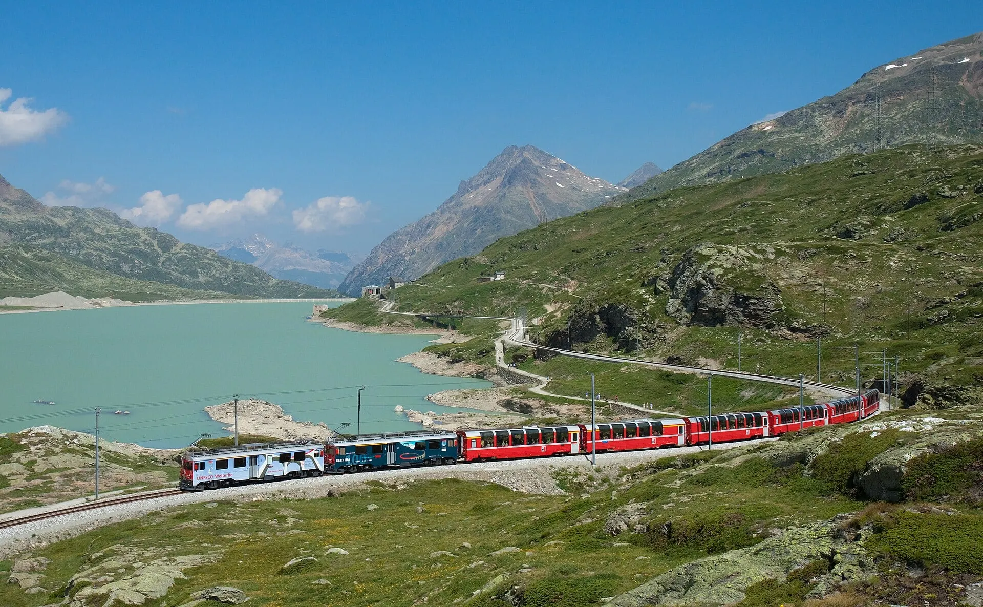 The Bernina Express passing the turquoise Lago Bianco under a blue sky, with Ospizio Bernina station in the background.