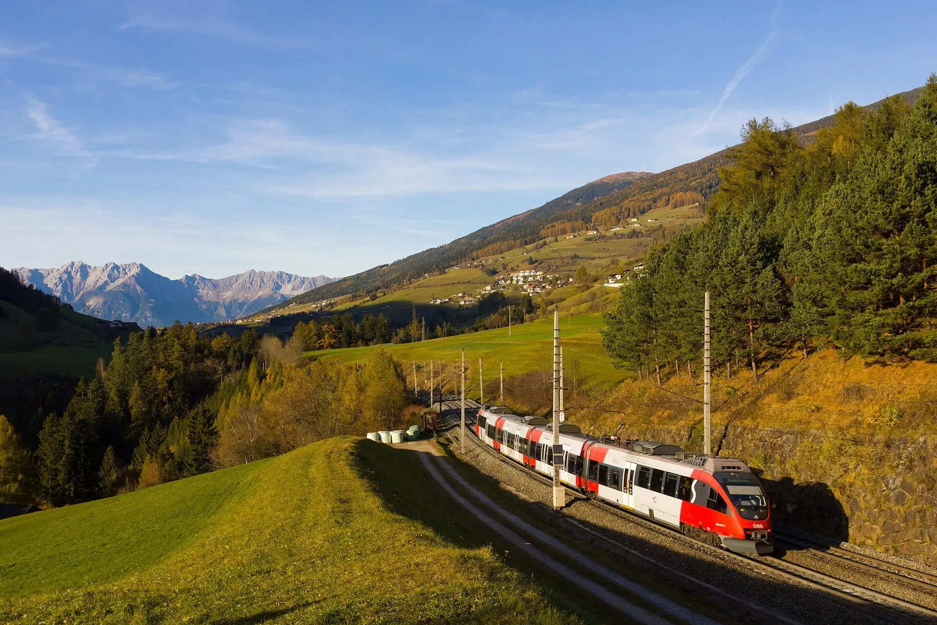 A red and white ÖBB train on the Brenner railway near Matrei, with autumn larches and mountain peaks under a blue sky
