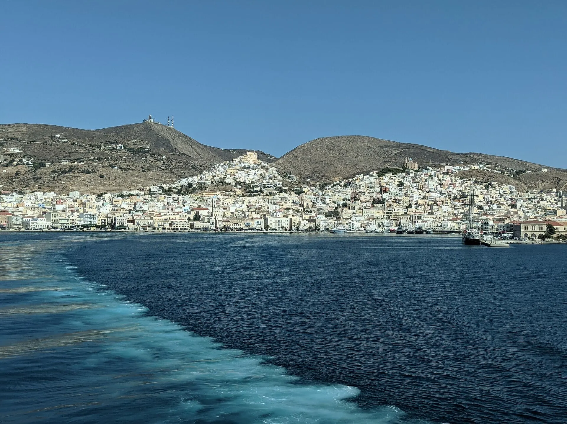 Die Zwillingshügel von Ermoupoli auf Syros von einer Fähre aus, deren Kielwasser sich über das Meer zieht