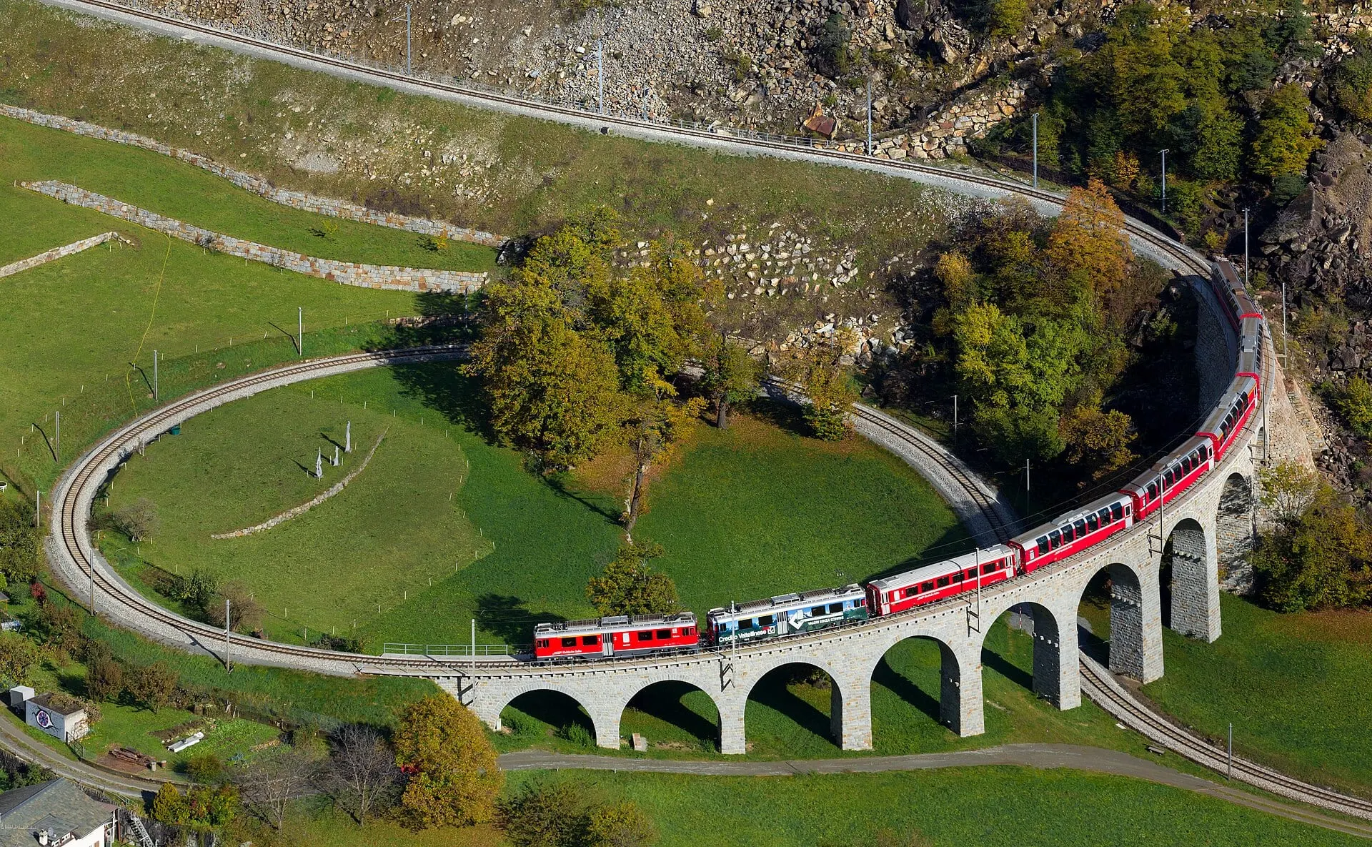 A red Rhaetian Railway train crossing the Brusio spiral viaduct, with the track looping round below it.