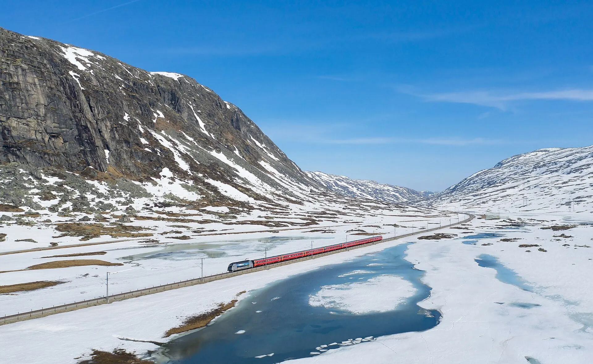 A Vy train crosses the snowy plateau above the tree line between Haugastøl and Finse, beside a half frozen lake.