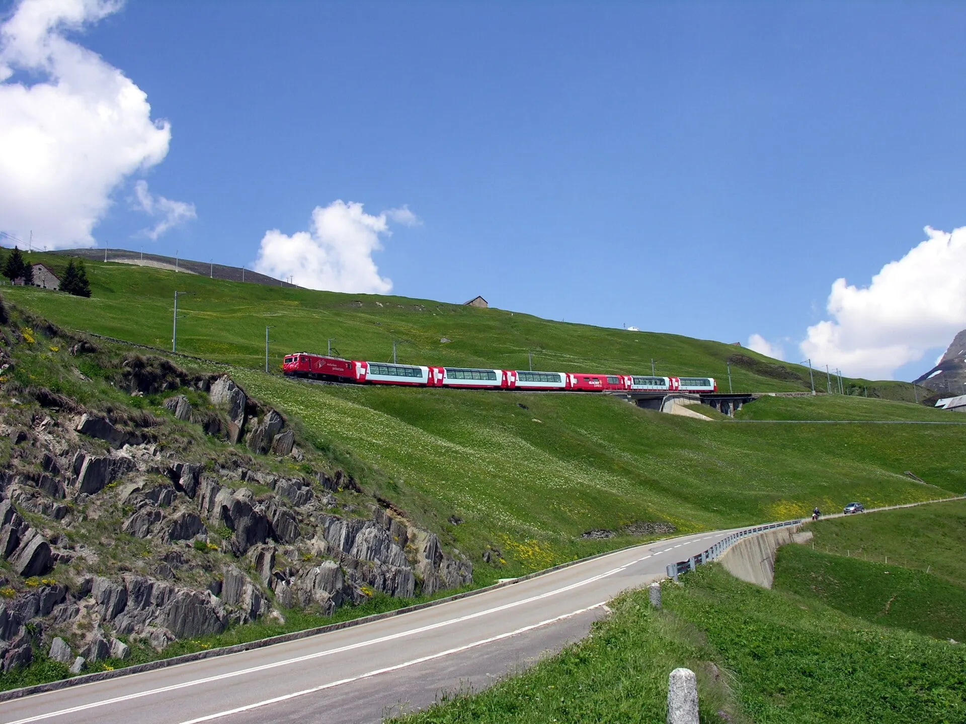 The Glacier Express climbing across green slopes towards the Oberalp Pass under a blue sky