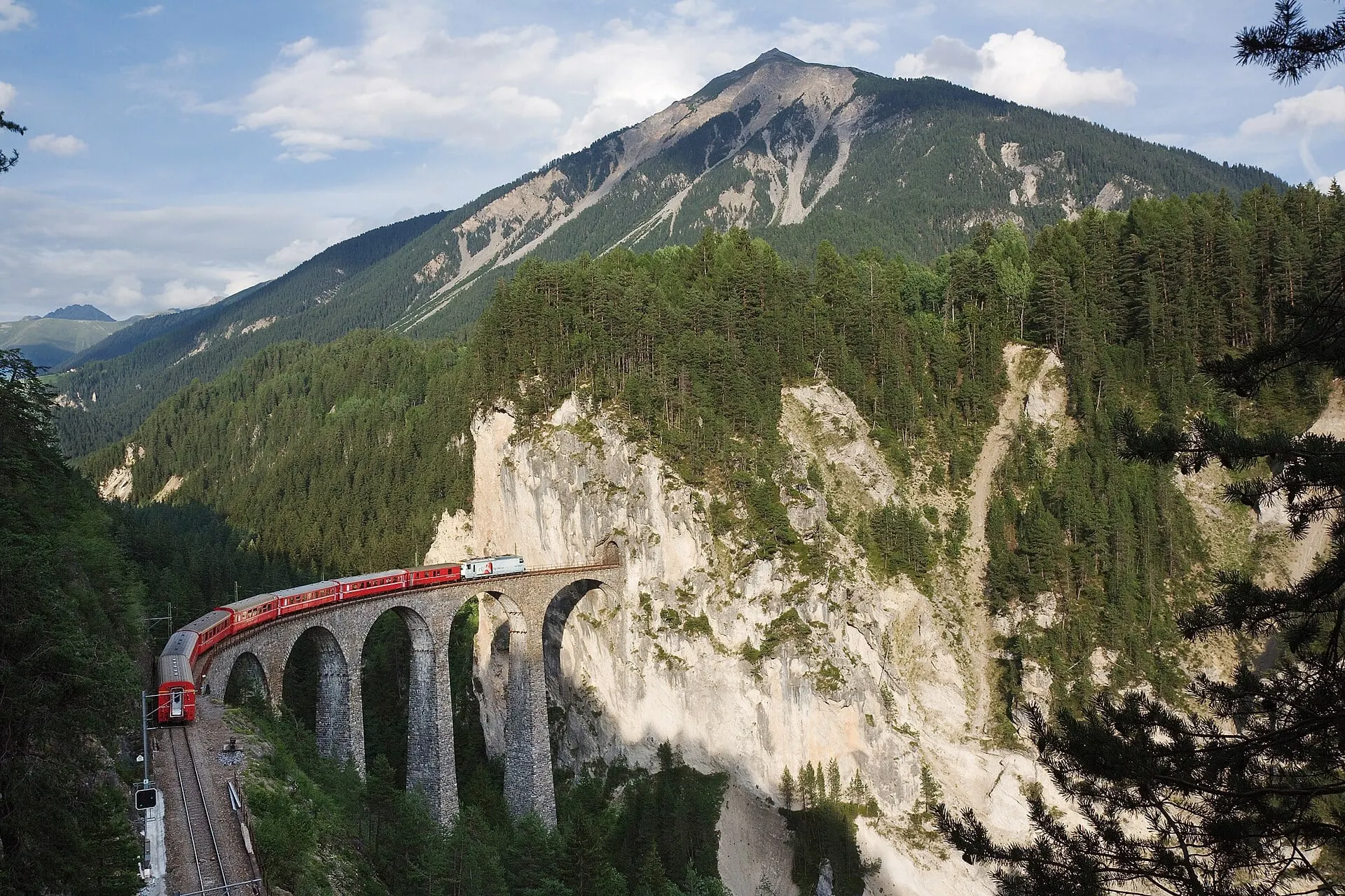 A red Rhaetian Railway train on the Landwasser Viaduct, running towards the tunnel in the cliff face.