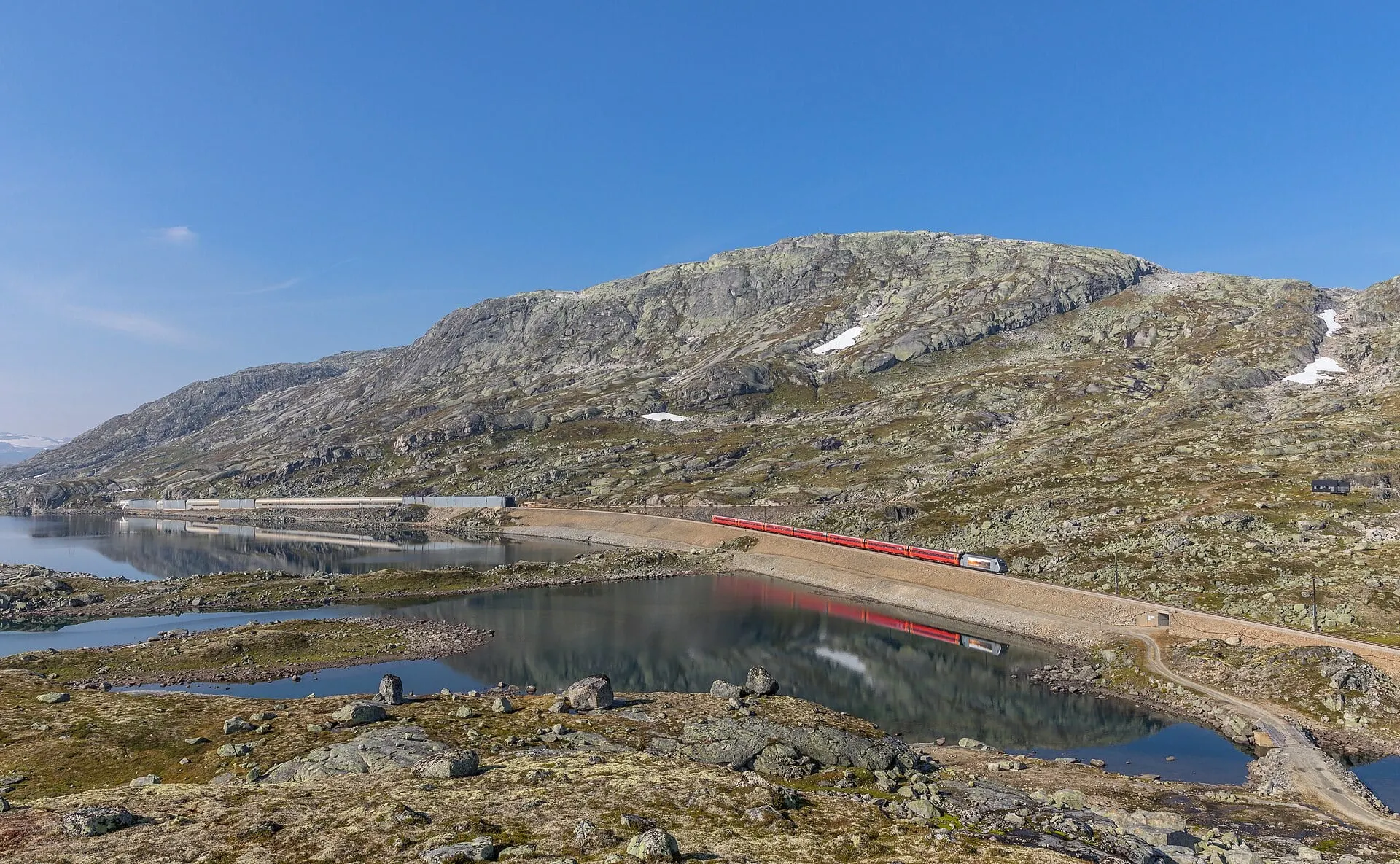 A train to Oslo runs beside a mountain lake between Hallingskeid and Finse, with the snow sheds of the old line on the far shore.