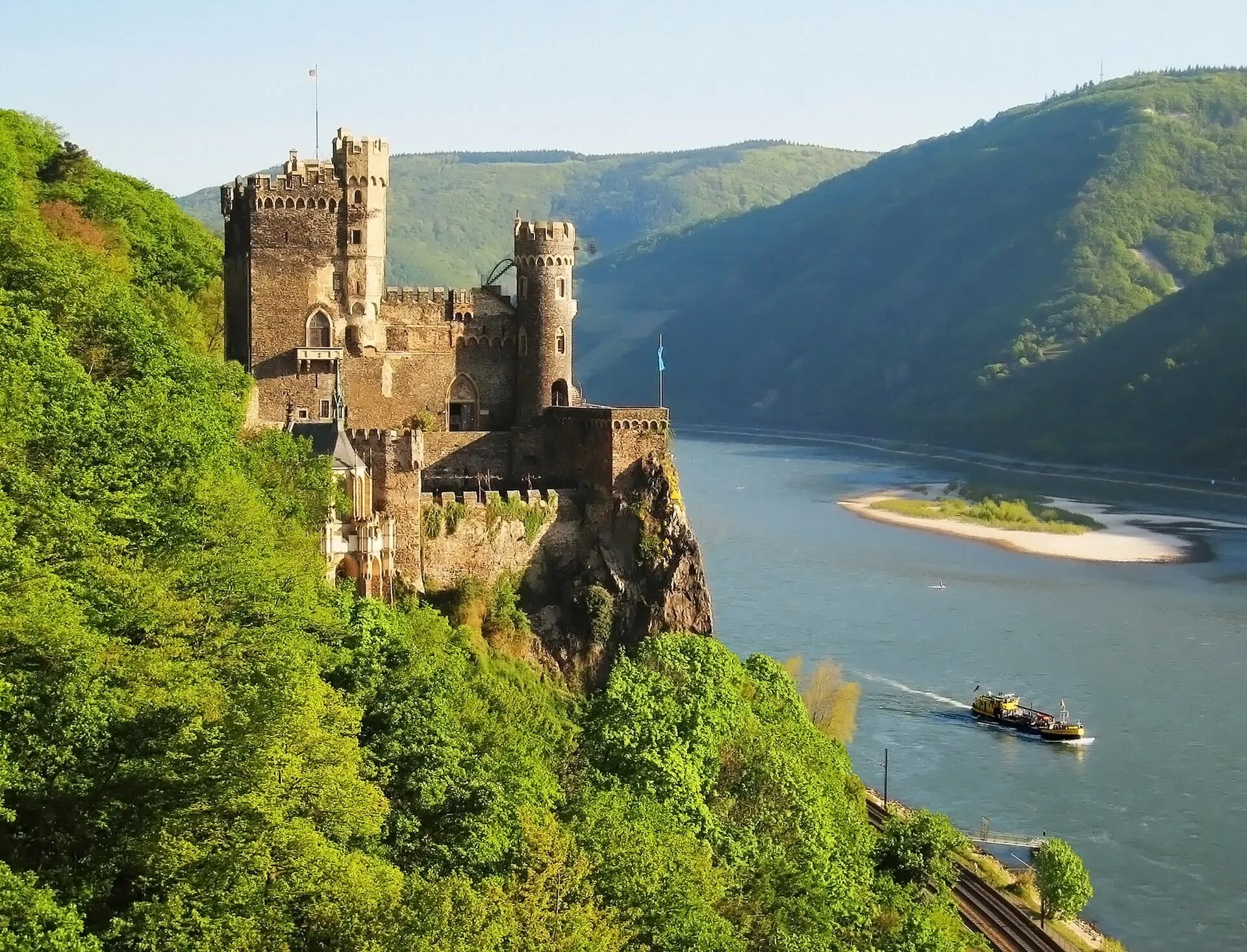 Rheinstein castle on its crag above the Rhine, with a barge on the river below.
