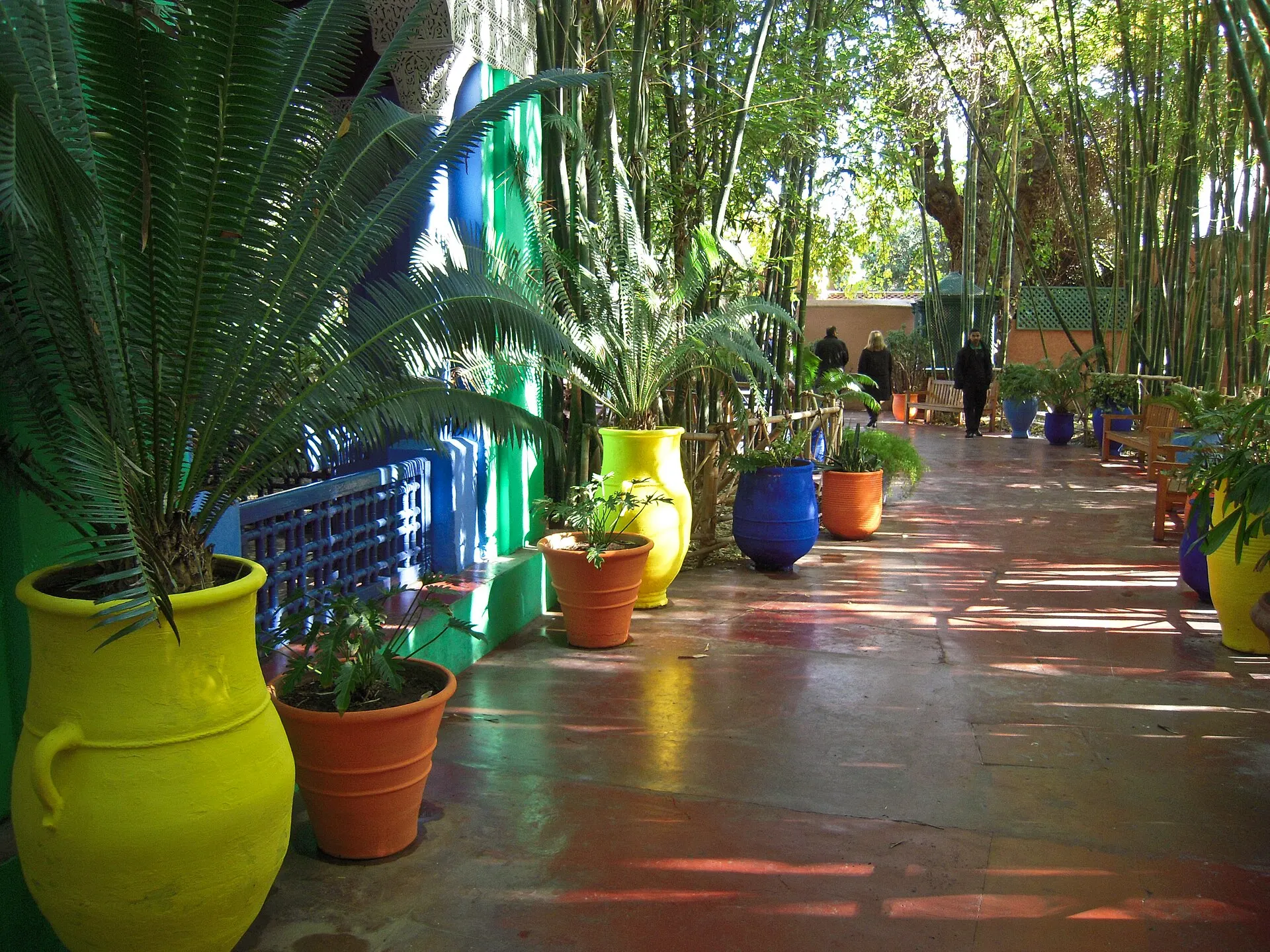A shaded walkway in the Jardin Majorelle lined with yellow, blue and terracotta pots and bamboo, with visitors in the distance