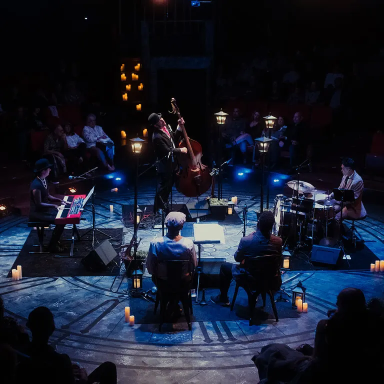 A Jazz Room band of piano, double bass, drums and horns playing on a round stage lit by lanterns, with the audience seated around them