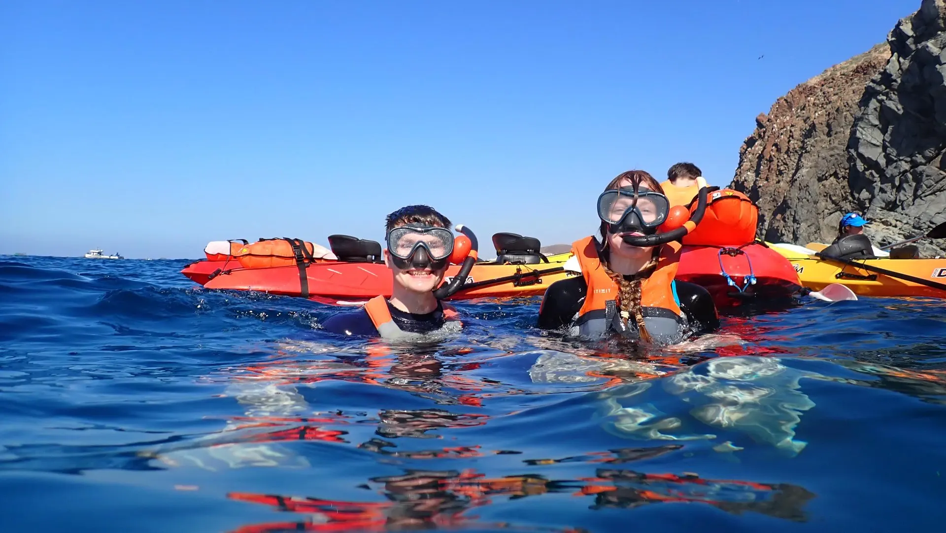 Snorkellers in masks floating beside a row of kayaks under a cliff