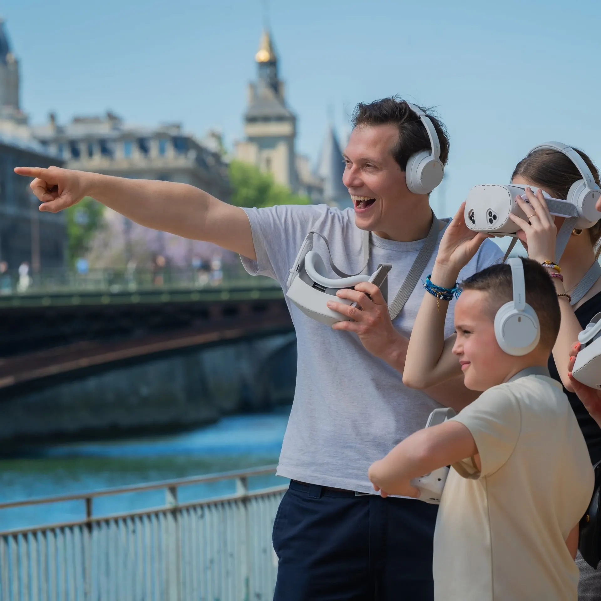 A man points across the Seine while a boy and a woman beside him hold white virtual reality viewers, all wearing headphones, on the right-bank quay with a bridge and the Conciergerie towers behind